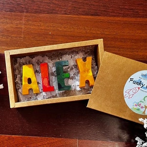 Może przedstawiać: Brązowe pudełko z przezroczystą pokrywką, w którym znajdują się kolorowe kredki w kształcie liter tworzących imię "ALEX". Pudełko jest oznaczone napisem "Funky Crayons" i zawiera grafikę rakiety i słońca.