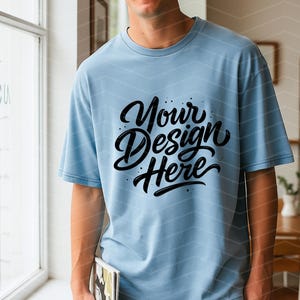 May include: Light blue t-shirt with the text "Your Design Here" in black script. The shirt is a solid color and has short sleeves. The person is holding a book.