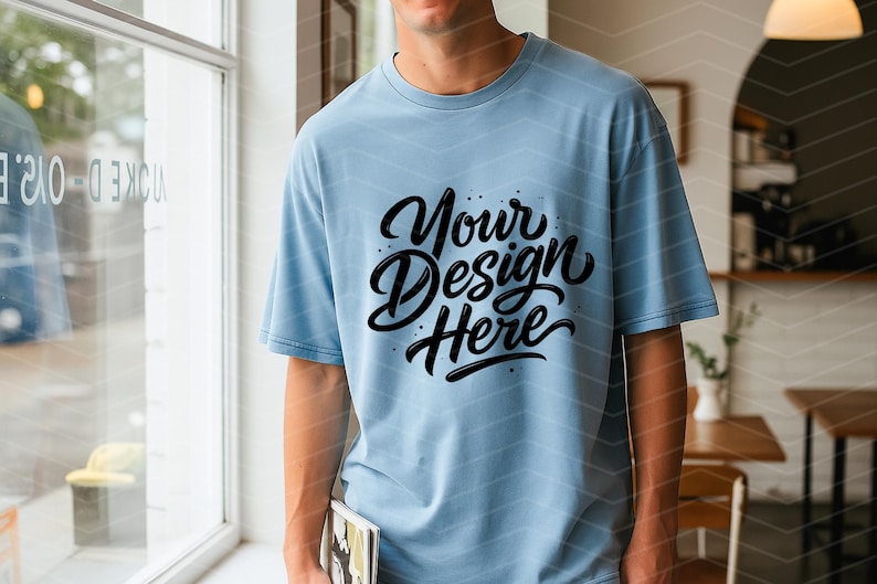 May include: Light blue t-shirt with the text "Your Design Here" in black script. The shirt is a solid color and has short sleeves. The person is holding a book.