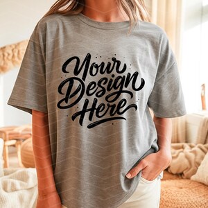 May include: A gray t-shirt with the text "Your Design Here" in a black, cursive font. The shirt is a relaxed fit and has short sleeves. The model is wearing white pants. The background is a cozy living room setting.