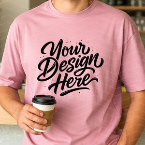 May include: A light pink t-shirt with the text "Your Design Here" in black script. The person is holding a white coffee cup with a brown sleeve. The background includes a wooden counter and a blurred interior.