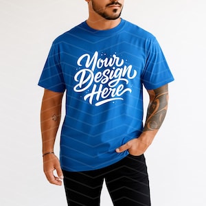 May include: A blue t-shirt with the text "Your Design Here" in white script. The shirt is short-sleeved and has a crew neck. The model is wearing black pants.