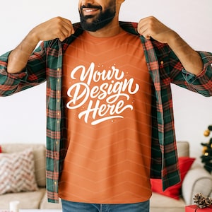 May include: Orange t-shirt with the text "Your Design Here" in white script. The shirt is worn under a green and red plaid flannel shirt. The person is smiling and wearing blue jeans.