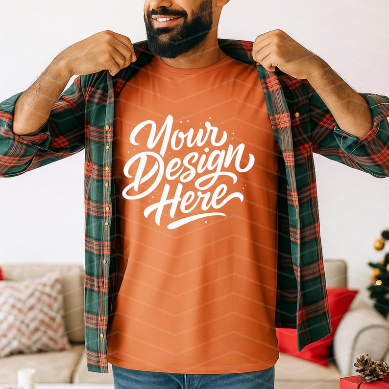 May include: Orange t-shirt with the text "Your Design Here" in white script. The shirt is worn under a green and red plaid flannel shirt. The person is smiling and wearing blue jeans.