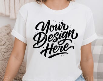 Puede incluir: Camiseta blanca con mangas enrolladas y el texto "Your Design Here" en negro, con una fuente cursiva. La camiseta es de estilo cuello redondo clásico, perfecta para mostrar diseños personalizados. El modelo lleva vaqueros azules.