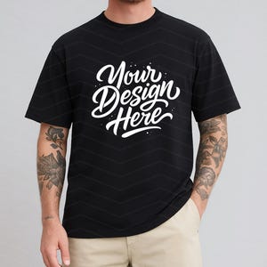 May include: Black t-shirt with the text "Your Design Here" in white script. The shirt has a crew neck and short sleeves. The model is wearing khaki pants.