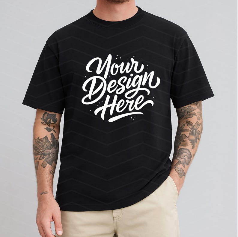 May include: Black t-shirt with the text "Your Design Here" in white script. The shirt has a crew neck and short sleeves. The model is wearing khaki pants.