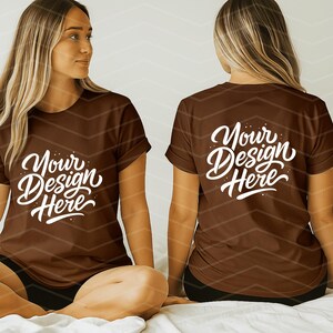 May include: Two brown t-shirts with the text "Your Design Here" in white script. The shirts are on a person sitting on a bed. The person is wearing black shorts. The background is a light color with a chevron pattern.