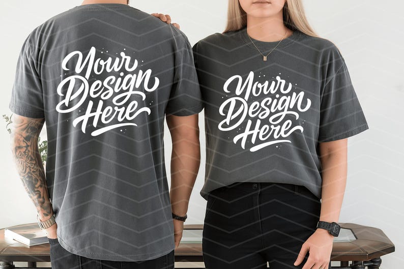 May include: Two charcoal gray t-shirts with the text "Your Design Here" in white script. The shirts are worn by two people. The background has a chevron pattern.