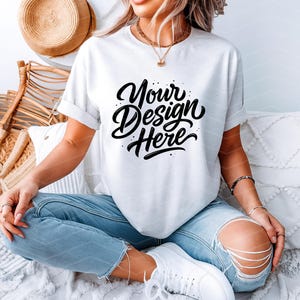 May include: White t-shirt with rolled sleeves featuring the text "Your Design Here" in a stylish, black script. The model is wearing light blue ripped jeans and white sneakers. A straw hat and chair are in the background.