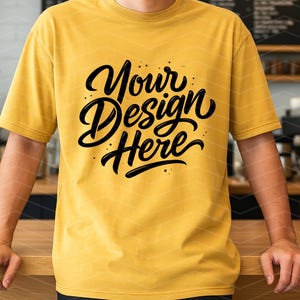 May include: Mustard yellow t-shirt with the text "Your Design Here" in a black, stylized script. The shirt is short-sleeved and worn by a person standing behind a wooden counter.