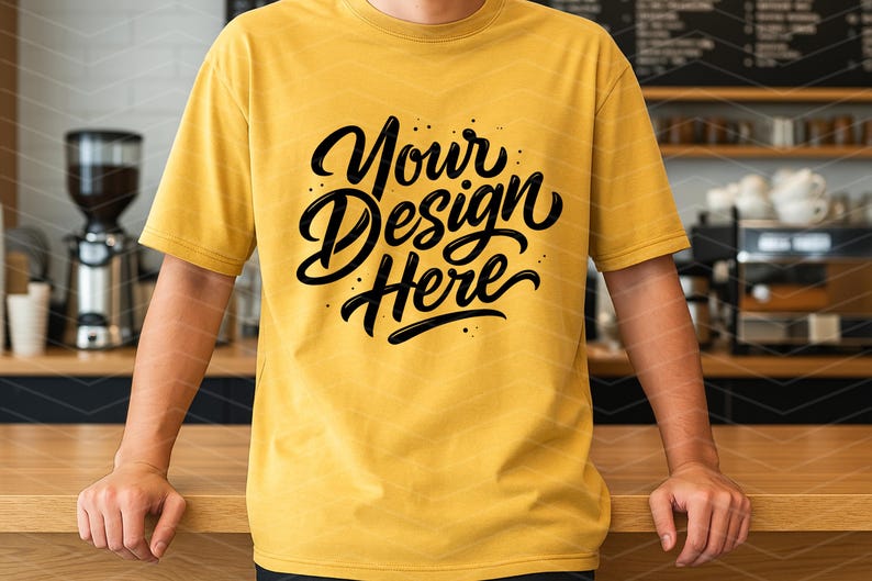 May include: Mustard yellow t-shirt with the text "Your Design Here" in a black, stylized script. The shirt is short-sleeved and worn by a person standing behind a wooden counter.