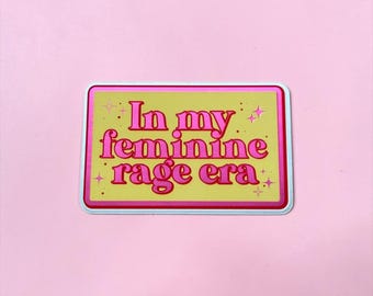 Pode incluir: Um autocolante retangular com fundo amarelo e rebordo cor-de-rosa. O texto In my feminine rage era está impresso numa fonte cor-de-rosa a negrito. Pequenos ícones de estrelas cor-de-rosa estão espalhados pelo texto centrado.