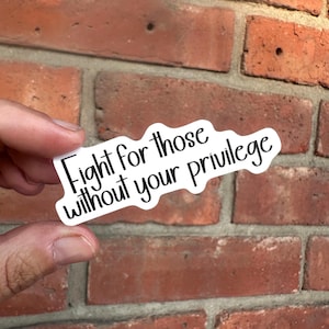 May include: A white sticker with black text that reads "Fight for those without your privilege." The sticker is held in front of a red brick wall.