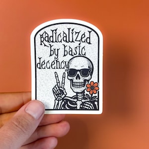 Puede incluir: Una pegatina blanca con una ilustración en negro de un esqueleto con gafas de sol y una flor, haciendo el signo de la paz. La pegatina tiene el texto "radicalized by basic decency" en una fuente estilizada.