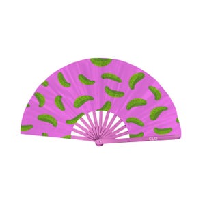 May include: A pink hand fan with a repeating pattern of green pickles. The fan is open, displaying the pickle design across its surface. The handle and frame are a matching pink color. The letters "CLQ" are printed on the bottom right.