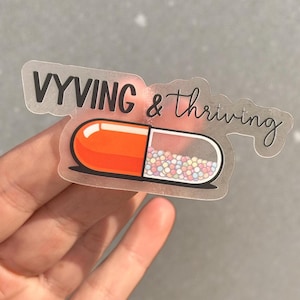 Può includere: Un adesivo trasparente con le parole "VYVING & thriving" in corsivo nero. L'adesivo presenta un design a capsula, con un'estremità arancione e l'altra metà riempita di perline colorate.