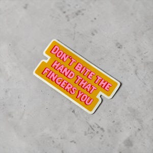 以下が含まれることがあります： ピンク色の「DON'T BITE THE HAND THAT FINGERS YOU」というテキストが入った黄色のステッカー。ステッカーには白い縁取りがあり、灰色の表面にあります。