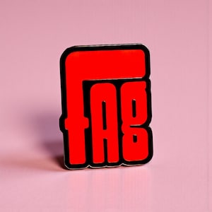 May include: A red enamel pin with the letters "FAG" in a bold, stylized font. The letters are outlined in black, and the pin is set against a pink background. The pin is a statement piece.