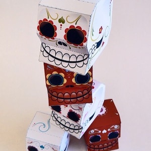 May include: A stack of four papercraft sugar skull boxes with colorful floral designs. The boxes are stacked on top of each other, with the top box being white and the bottom box being brown.