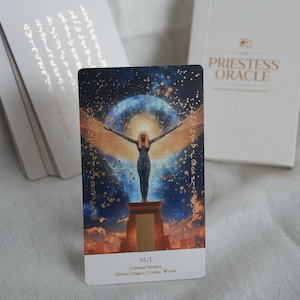 May include: A tarot card from "The Priestess Oracle" deck featuring an illustration of Nut, the celestial mother, with outstretched wings against a starry background. The card has the word "NUT" at the bottom, along with other text. A white box with the deck's name is in the background.