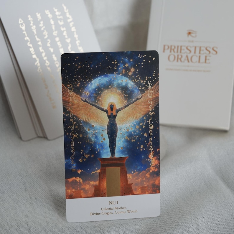 May include: A tarot card from "The Priestess Oracle" deck featuring an illustration of Nut, the celestial mother, with outstretched wings against a starry background. The card has the word "NUT" at the bottom, along with other text. A white box with the deck's name is in the background.