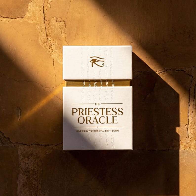 May include: A white rectangular deck box titled The Priestess Oracle Divine Light Codes of Ancient Egypt. The box features a gold Eye of Horus symbol at the top and gold lettering on a white background with subtle embossed hieroglyphic patterns.