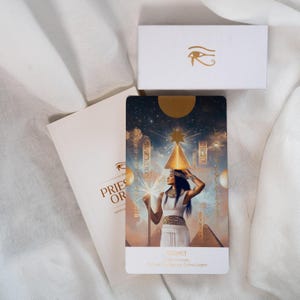 May include: A rectangular tarot card featuring an illustration of a figure in white robes holding a staff beneath a golden pyramid, with the text SES HET. Beside it is a white booklet titled THE PRIESTESS ORACLE and a small white box with a gold eye symbol.