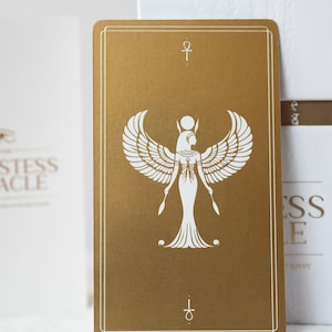 May include: A gold-colored tarot card featuring a white illustration of a winged figure with a headdress, holding two scepters. The card has a white border and an ankh symbol at the top and bottom. The card is part of a deck.