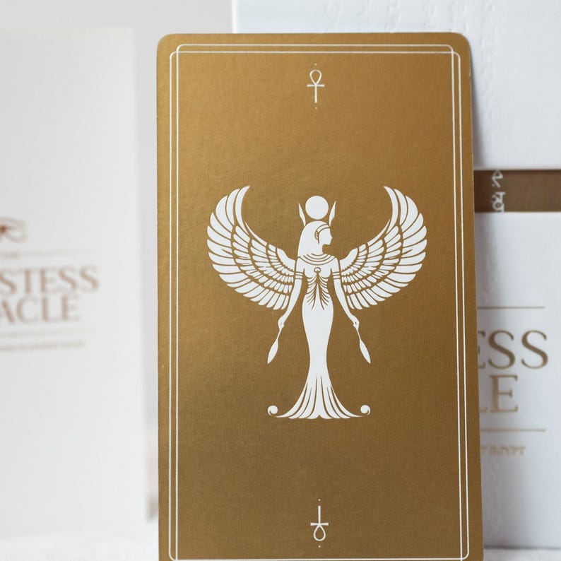 May include: A gold-colored tarot card featuring a white illustration of a winged figure with a headdress, holding two scepters. The card has a white border and an ankh symbol at the top and bottom. The card is part of a deck.