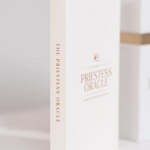 May include: A white box with the words "THE PRIESTESS ORACLE" printed vertically in a gold color on the spine. The front of the box has the words "PRIESTESS ORACLE" in gold, with a small symbol above the text.
