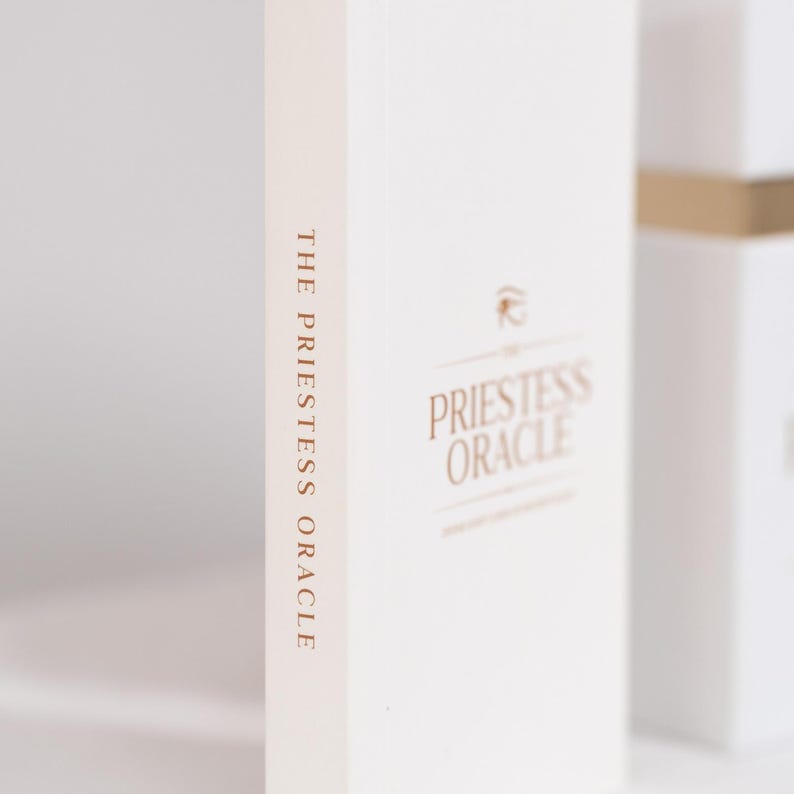 May include: A white box with the words "THE PRIESTESS ORACLE" printed vertically in a gold color on the spine. The front of the box has the words "PRIESTESS ORACLE" in gold, with a small symbol above the text.