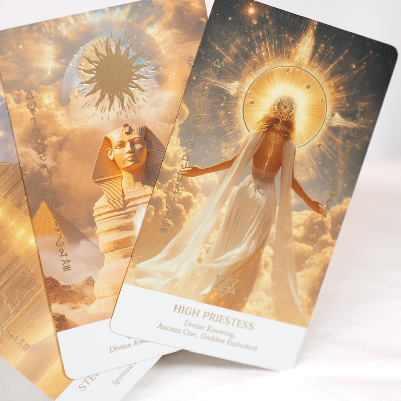 May include: Two rectangular tarot cards with gold-toned imagery. One card features a sphinx and pyramid under a sun, while the other depicts a figure in a white gown with a glowing halo, labeled HIGH PRIESTESS, Divine Knowing, Ancient One, Goddess Embodied.