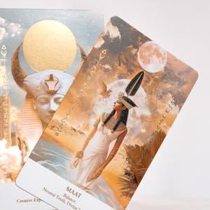 May include: Two oracle cards with Egyptian-themed illustrations. One card features a woman in a white dress and headdress, with the word "MAAT" and related concepts. The other card shows a pharaoh with a golden sun disc. The cards have a warm color palette.