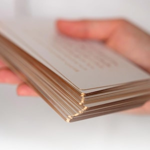 May include: A stack of rectangular white cards with metallic gold gilded edges. The cards are held in a fan-like arrangement, showcasing the reflective gold finish on the sides of each individual card against the clean white paper surfaces.