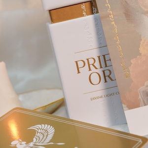 May include: A white rectangular box with the words "PRIE OR" in gold lettering. The box also has the text "DIVINE LIGHT CO" and gold hieroglyphic-style text. A gold-colored card with an illustration is in the foreground.
