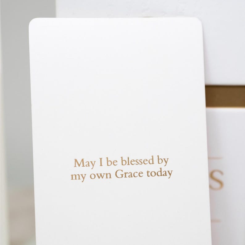 May include: A rectangular white card with rounded corners featuring the gold-colored text May I be blessed by my own Grace today centered on the lower half of the surface. The card has a smooth matte finish and a clean, simple design.