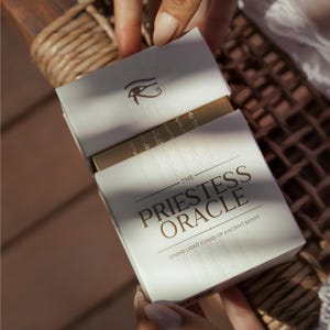 May include: A white box with gold lettering and the text "THE PRIESTESS ORACLE DIVINE LIGHT CODES OF ANCIENT EGYPT" is opened to reveal a gold-colored deck of cards. The box has an embossed eye symbol.