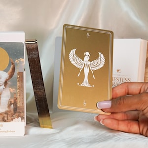 May include: A gold-colored tarot card featuring a white illustration of a winged figure, held in a hand. The card has an ankh symbol at the top and bottom. Other cards and a box are visible in the background.