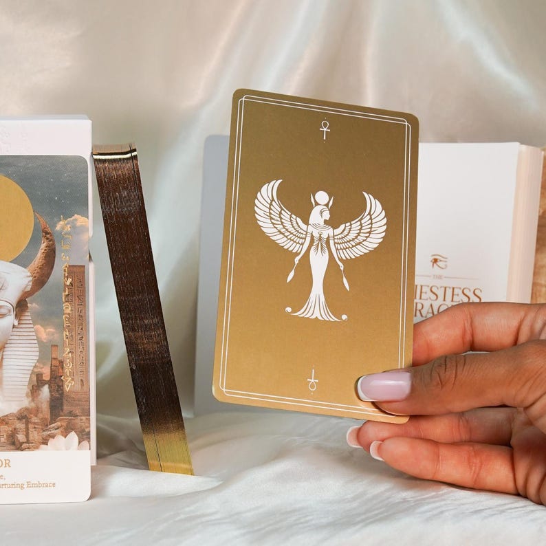 May include: A gold-colored tarot card featuring a white illustration of a winged figure, held in a hand. The card has an ankh symbol at the top and bottom. Other cards and a box are visible in the background.