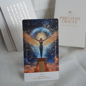 May include: A tarot card from the Priestess Oracle deck featuring an illustration of Nut, the celestial mother, with outstretched wings against a starry background. The card is predominantly blue, gold, and black, with the word "NUT" at the bottom.