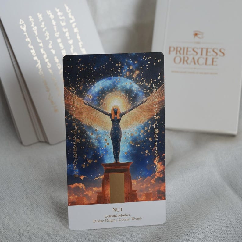 May include: A tarot card from the Priestess Oracle deck featuring an illustration of Nut, the celestial mother, with outstretched wings against a starry background. The card is predominantly blue, gold, and black, with the word "NUT" at the bottom.