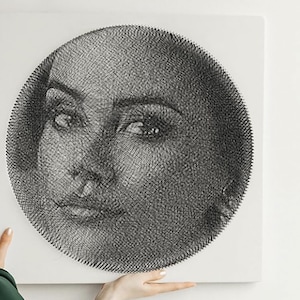 May include: A black and white string art portrait of a person's face, set against a white background. The artwork is circular, with the face centered and detailed, showcasing the eyes, nose, and lips. The string art creates a textured effect.