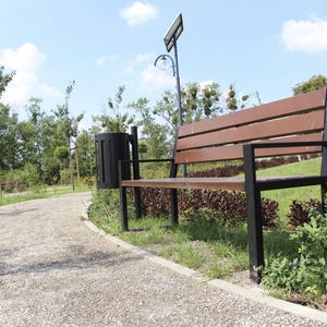 Könnte beinhalten: Eine Holzbank mit schwarzem Metallrahmen steht auf einem Schotterweg in einem Park. Die Bank ist von grünem Gras und Bäumen umgeben.