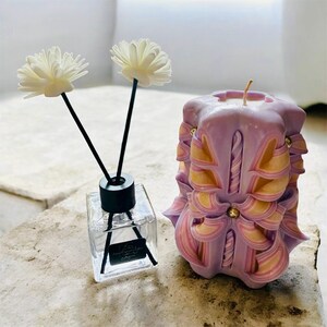 May include: A decorative arrangement featuring a clear glass diffuser with two white flower reeds and a carved purple candle with yellow and pink accents. The candle has a bow design and a gold bead.