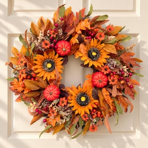 May include: A circular autumn wreath featuring artificial sunflowers with dark centers, small red pumpkins, clusters of red berries, and a variety of orange, yellow, and brown maple leaves. The arrangement is dense and textured with foliage.