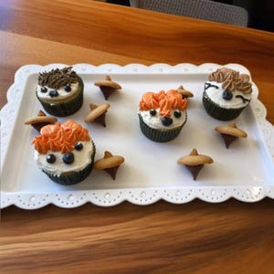 May include: A white platter with four decorated cupcakes and six chocolate acorns. The cupcakes are topped with orange and brown frosting and have animal faces.