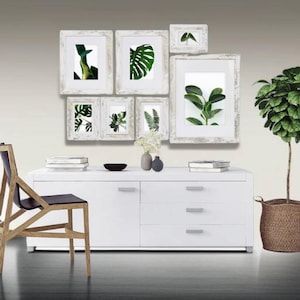May include: A white dresser with four drawers and a white top. The dresser is decorated with a collection of white picture frames with green leaf prints. A potted plant with green leaves is on the right side of the dresser.