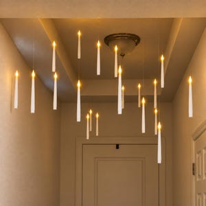 Può includere: Una collezione di candele coniche bianche a LED sospese al soffitto a diverse altezze. Ogni candela presenta una fiamma gialla calda e luminosa sulla punta, creando un effetto fluttuante in tutto lo spazio del corridoio vicino a una plafoniera.