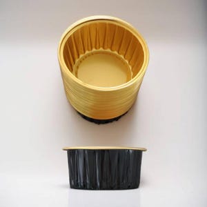 May include: A stack of gold-colored aluminum baking cups, with a single black aluminum baking cup in the foreground.